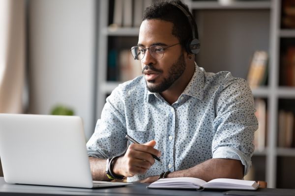 Person wearing a headset working on a laptop while taking notes at a desk.

